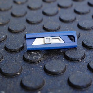 May include: A blue utility knife with a silver blade and a gray slider. The knife is resting on a textured, dark gray surface with a pattern of raised, rounded shapes. The blade is partially exposed.