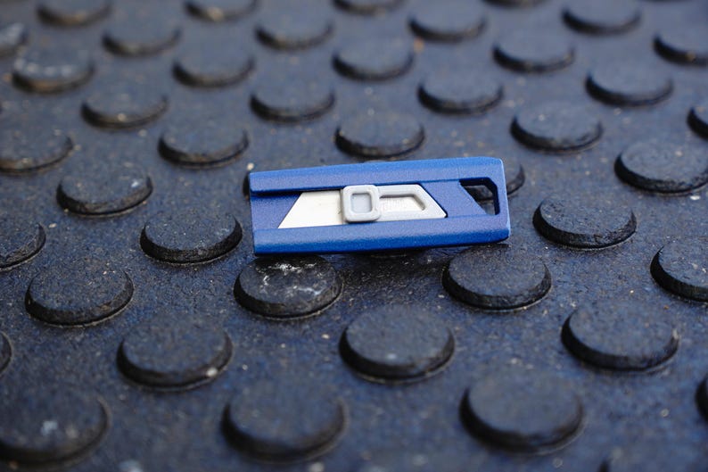 May include: A blue utility knife with a silver blade and a gray slider. The knife is resting on a textured, dark gray surface with a pattern of raised, rounded shapes. The blade is partially exposed.