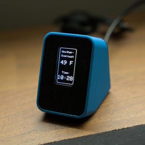 May include: A teal-colored, triangular-shaped device with a black screen displaying weather information and time. The screen shows "Overcast" weather, a temperature of 49°F, and the time 10:28. The device is on a wooden surface.