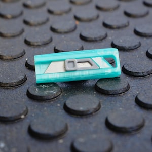 May include: A teal utility knife with a silver blade and gray accents rests on a textured black surface. The knife has a rectangular shape with a hole at one end. The blade is partially exposed.