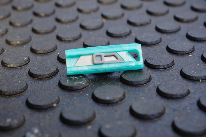 May include: A teal utility knife with a silver blade and gray accents rests on a textured black surface. The knife has a rectangular shape with a hole at one end. The blade is partially exposed.