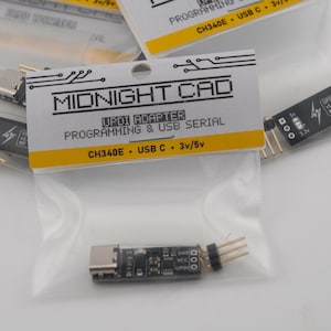 May include: A small black electronic UPDI adapter with a USB-C connector and three gold pins, packaged in a clear plastic bag with a white header card labeled Midnight Cad, UPDI Adapter, Programming & USB Serial, CH340E, USB C, 3v/5v.