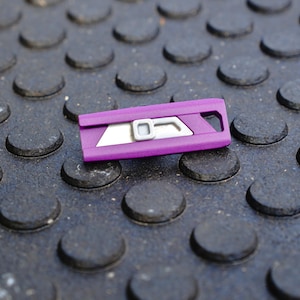 May include: A purple utility knife with a silver blade and accents rests on a textured, dark gray surface. The knife has a rectangular shape with a curved handle and a small opening at one end.