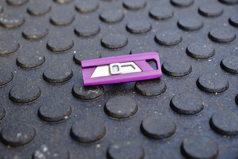 May include: A purple utility knife with a silver blade and accents rests on a textured, dark gray surface. The knife has a rectangular shape with a curved handle and a small opening at one end.