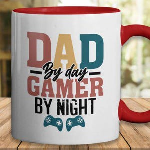 May include: White ceramic mug with a red handle and interior. The mug features the text "DAD By day GAMER By night" in various colors, along with two teal game controller icons. A fun gift for gamers.