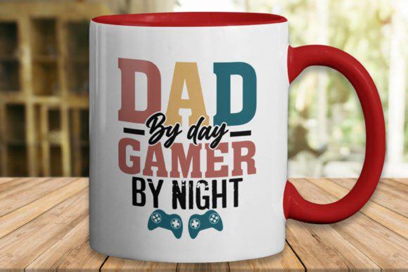 May include: White ceramic mug with a red handle and interior. The mug features the text "DAD By day GAMER By night" in various colors, along with two teal game controller icons. A fun gift for gamers.