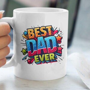 May include: White ceramic mug with the text "BEST DAD EVER" in a colorful, comic book style design. The mug is held by a person's hand. A croissant rests on a napkin in the background.