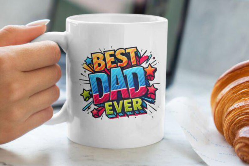 May include: White ceramic mug with the text "BEST DAD EVER" in a colorful, comic book style design. The mug is held by a person's hand. A croissant rests on a napkin in the background.