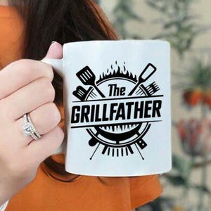 May include: White ceramic mug with the text "The GRILLFATHER" in black, featuring grilling tools and flames. A perfect gift for the grill master. The mug is held by a person.