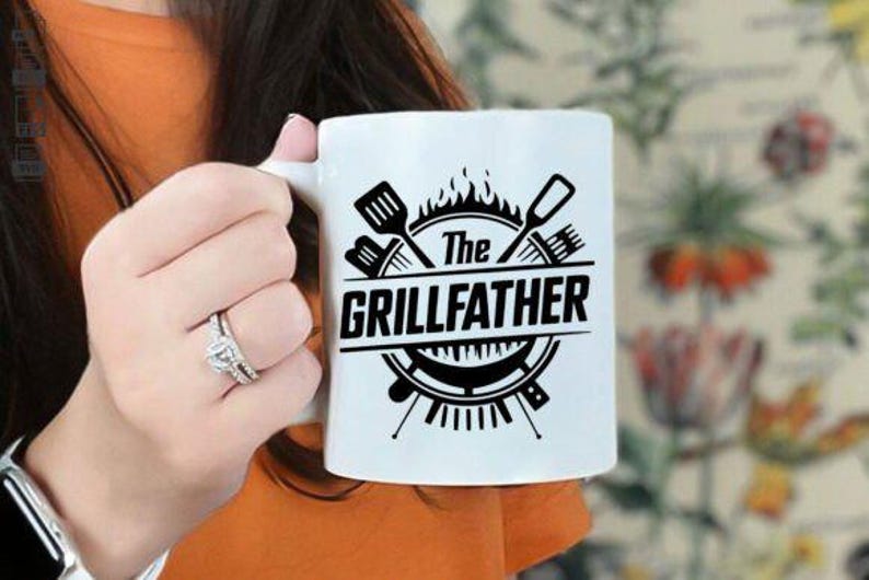 May include: White ceramic mug with the text "The GRILLFATHER" in black, featuring grilling tools and flames. A perfect gift for the grill master. The mug is held by a person.