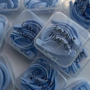 May include: Several clear plastic containers filled with light blue lash lift tools. Each container has the words "LAMI TIME" printed on it in black. The tools are arranged in a curved shape.