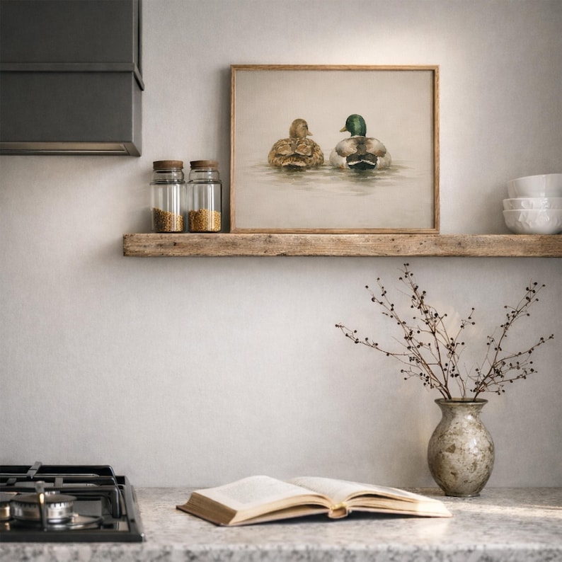 Pu&ograve; includere: Stampa incorniciata con due anatre sull'acqua, con dettagli di piume. L'opera &egrave; esposta su una mensola di legno rustico. L'ambiente cucina include barattoli, un vaso con rami e un libro aperto, creando un'atmosfera naturale.