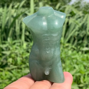 Peut inclure: Une figurine de torse sculptée, vert jade translucide. La sculpture représente une forme masculine, avec une musculature définie et des détails anatomiques. La figurine est tenue dans une main sur fond de feuillage vert.