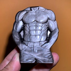 Peut inclure: Une sculpture de torse sans tête grise représentant un physique musclé. La sculpture présente une définition détaillée de la poitrine, de l'abdomen et des bras, avec les mains dans les poches du pantalon. La figurine est dans une pose détendue.