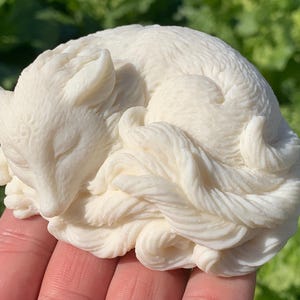 May include: A white ceramic sculpture of a fox curled up sleeping. The sculpture is detailed with fur and has a realistic appearance.