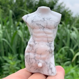 Peut inclure: Une sculpture de torse en marbre blanc avec des veines grises. La sculpture représente une figure masculine avec des muscles abdominaux définis. La sculpture est tenue dans une main sur fond de feuillage vert.