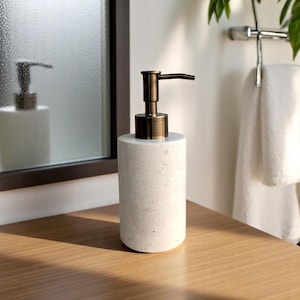 May include: A cylindrical soap dispenser with a textured, light grey stone base and a bronze-coloured pump. The dispenser is on a wooden surface, with a mirror and a towel rail in the background. The design is minimalist and modern.