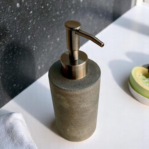 Könnte beinhalten: Ein zylindrischer Seifenspender mit einem Pumpenkopf aus gebürstetem Messing. Der Körper hat eine strukturierte, graubraune Farbe. Der Spender steht auf einer weißen Oberfläche, mit einem weißen Handtuch und einem dekorativen Gegenstand im Hintergrund.