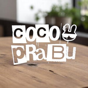 May include: White cutout letters spelling "COCO PraBu" with a cartoon rabbit wearing sunglasses. The letters are arranged in a layered design, with some appearing to drip. The cutout is on a wooden surface.