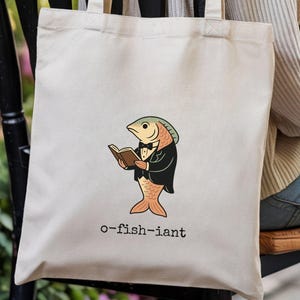 May include: Beige tote bag featuring a cartoon fish in a tuxedo reading a book. The fish is orange, with a green and gold head, and a black tuxedo. The text "o-fish-iant" is printed below the fish.