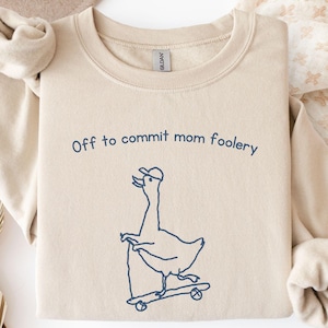 May include: A sand-colored crewneck sweatshirt featuring a dark blue line art graphic of a duck wearing a baseball cap while riding a skateboard. The text above the duck reads, "Off to commit mom foolery" in a simple, dark blue sans-serif font.