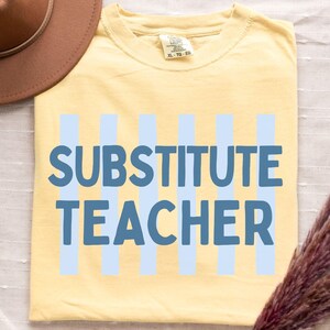 May include: A pale yellow t-shirt featuring the text SUBSTITUTE TEACHER in bold blue letters over vertical light blue stripes. The shirt is displayed alongside a brown wide-brimmed hat and a bundle of dried pampas grass, with the word BUTTER printed below.