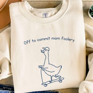 Peut inclure: Sweat-shirt crème avec le texte "Off to commit mom foolery" et un dessin d'oie sur un skateboard. L'image comprend un chapeau marron, un jean bleu, des sandales et une petite plante succulente.