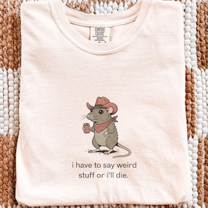 May include: Ivory-colored t-shirt with a graphic of a cartoon rat wearing a cowboy hat and holding a coffee cup. The text "i have to say weird stuff or i'll die." is printed below the graphic. The shirt is made of a soft material.