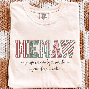 Könnte beinhalten: Elfenbeinfarbenes T-Shirt mit dem Wort "MEMAW" in einer dekorativen Schriftart, mit roten, grünen und herzförmigen Mustern. Unter dem Wort stehen die Namen "jasper", "emily", "sarah", "jennifer" und "noah" in Schreibschrift.
