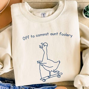 Puede incluir: Sudadera color crema con la frase "Off to commit aunt foolery" y un dibujo de un ganso en monopatín. La imagen incluye pantalones cortos de mezclilla azules, un sombrero beige y sandalias. También se ve una pequeña planta suculenta.