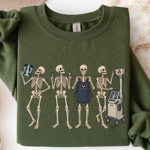 May include: A military green crewneck sweatshirt featuring a graphic print of four skeletons. One skeleton holds an X-ray, two hold markers labeled L and R, and one wears a blue apron with a radiation symbol while operating an X-ray machine.