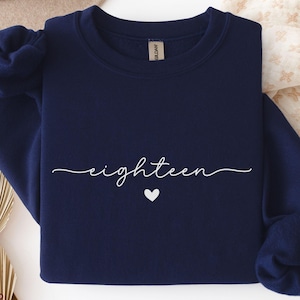 May include: Navy blue sweatshirt with the word "eighteen" in a cursive font and a small white heart underneath. The sweatshirt is a crew neck style and has long sleeves. The word "NAVY" is printed in a tan font at the bottom left.
