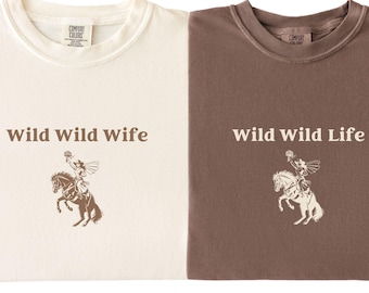 May include: Two t-shirts, one ivory and one espresso, with the text "Wild Wild Wife" and "Wild Wild Life". Each shirt features a graphic of a person riding a horse. The text "IVORY & ESPRESSO" is at the bottom.