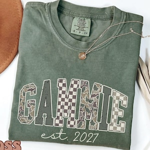Op de afbeelding: Een mosgroen t-shirt met korte mouwen, een ronde hals en een Comfort Colors merklabel. Op de voorzijde staat de tekst GAMMIE in een groot, gebogen, verweerd lettertype met afwisselende camouflage- en ruitpatronen, gevolgd door est. 2027.