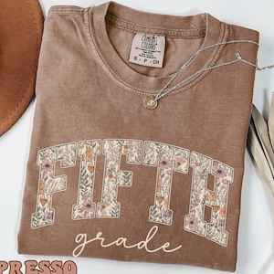 May include: A brown short-sleeved t-shirt with a crew neckline. The front features the word FIFTH in large block letters filled with a floral pattern, and the word grade written below in white cursive script. The shirt has a Comfort Colors label.