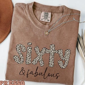 May include: Espresso-colored t-shirt with the words "Sixty & fabulous" in a leopard print and script font. The shirt has a crew neck and short sleeves. A silver necklace with a pendant is on the shirt.