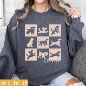 May include: Dark heather sweatshirt featuring a grid of nine dog illustrations in beige squares. The dogs are in various poses, with the name "Charlie" in cursive below. The sweatshirt is a dark gray color.
