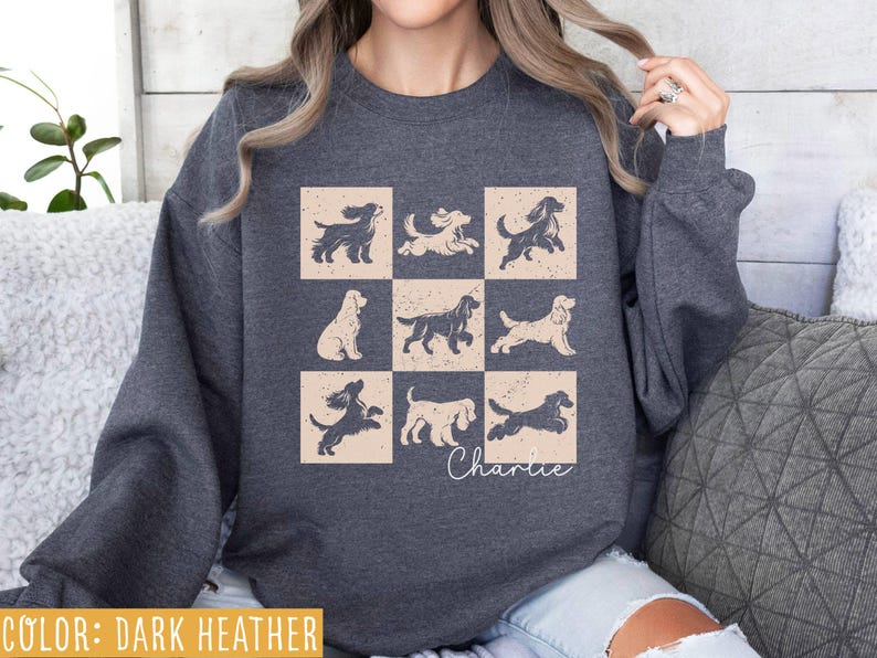 May include: Dark heather sweatshirt featuring a grid of nine dog illustrations in beige squares. The dogs are in various poses, with the name "Charlie" in cursive below. The sweatshirt is a dark gray color.