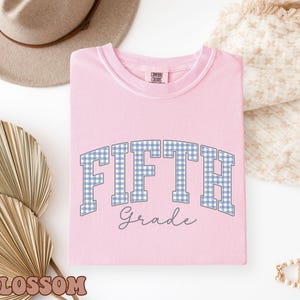 May include: A light pink short-sleeved t-shirt featuring the text FIFTH in a blue and white gingham pattern with the word Grade written below in a dark blue script font. The shirt has a Comfort Colors brand label on the inner neck collar.
