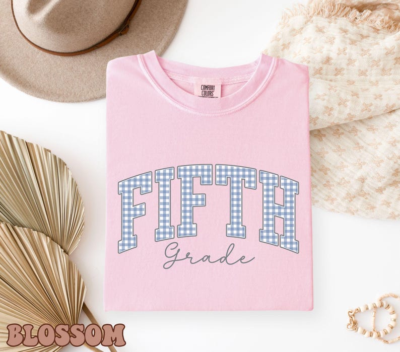 May include: A light pink short-sleeved t-shirt featuring the text FIFTH in a blue and white gingham pattern with the word Grade written below in a dark blue script font. The shirt has a Comfort Colors brand label on the inner neck collar.