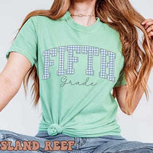 May include: A light green short-sleeve t-shirt featuring the word FIFTH in a blue and white gingham pattern with a dark outline, and the word Grade written in a cursive script font below it. The shirt is tied in a knot at the bottom hem.