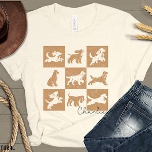 May include: Cream-colored t-shirt featuring a grid of nine dog illustrations in brown and white. The name "Charlie" is written in cursive below the grid. The shirt is paired with blue jeans and a brown cowboy hat.