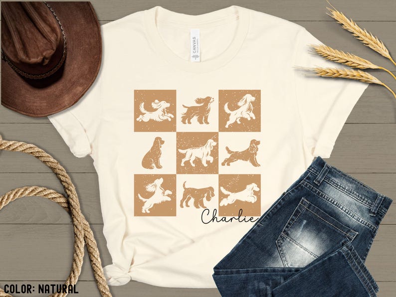 May include: Cream-colored t-shirt featuring a grid of nine dog illustrations in brown and white. The name "Charlie" is written in cursive below the grid. The shirt is paired with blue jeans and a brown cowboy hat.