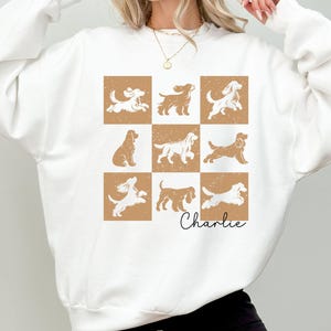 May include: White sweatshirt featuring a grid of nine dog illustrations in various poses, set against a tan background. The name "Charlie" is written in cursive below the grid. The word "WHITE" is printed at the bottom left.