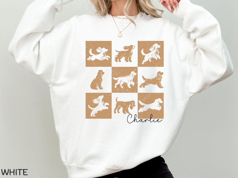 May include: White sweatshirt featuring a grid of nine dog illustrations in various poses, set against a tan background. The name "Charlie" is written in cursive below the grid. The word "WHITE" is printed at the bottom left.