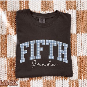 May include: A black crewneck t-shirt featuring the word FIFTH in a blue and white gingham block font, arched across the chest. Below the word FIFTH, the word Grade is printed in a white cursive script. The shirt has a standard collar.
