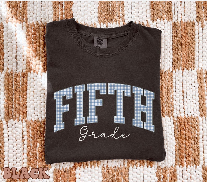 May include: A black crewneck t-shirt featuring the word FIFTH in a blue and white gingham block font, arched across the chest. Below the word FIFTH, the word Grade is printed in a white cursive script. The shirt has a standard collar.