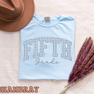 May include: A light blue t-shirt featuring the text FIFTH in a blue and white gingham pattern with the word Grade written in a cursive script below it. The shirt is accompanied by a brown wide-brimmed hat and a bundle of dried purple pampas grass.