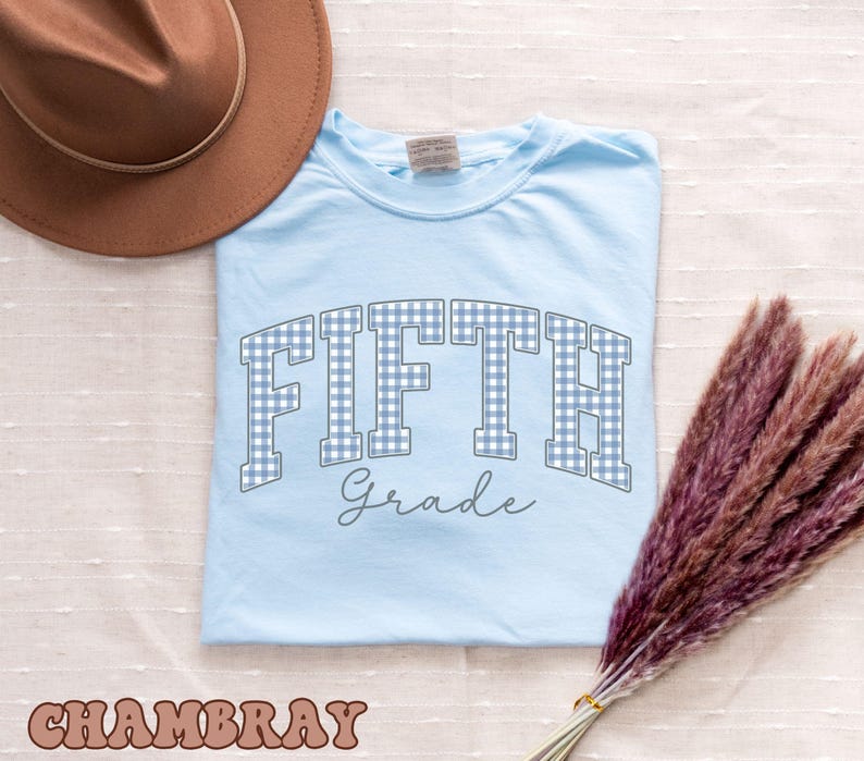 May include: A light blue t-shirt featuring the text FIFTH in a blue and white gingham pattern with the word Grade written in a cursive script below it. The shirt is accompanied by a brown wide-brimmed hat and a bundle of dried purple pampas grass.