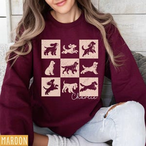 May include: Maroon sweatshirt featuring a grid of dog illustrations in various poses. The design includes the name "Charlie" in cursive. The sweatshirt is a solid maroon color, with a distressed, vintage-style graphic.